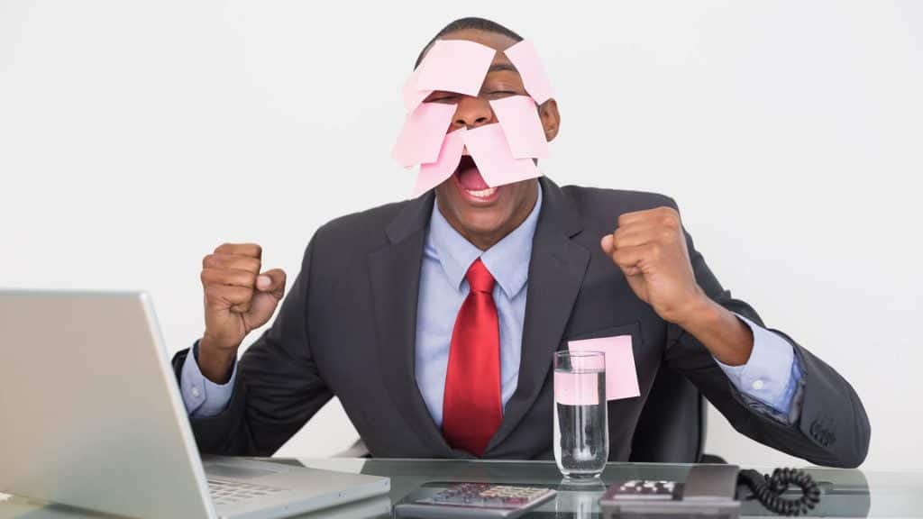 Black man wearing a suit and tie holding several pink sticky notes on his face. He screams desperately in front of a laptop.