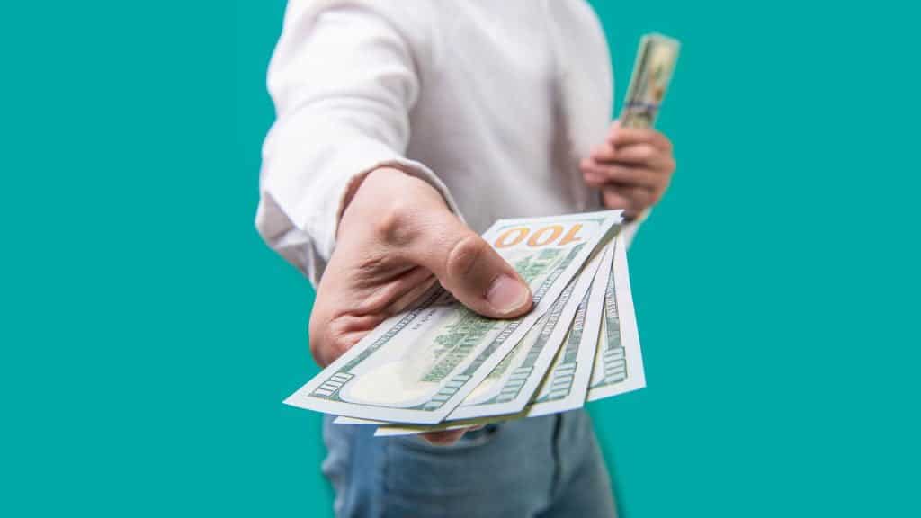Sectional photograph of a man standing in front of a light green background. The man's hand can be seen in front holding several $100 bills.