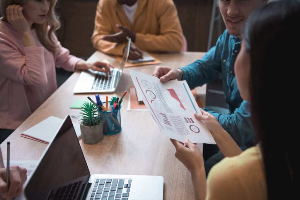 A group of young professionals discussing charts and graphs around a table, illustrating "conduct a business impact analysis."