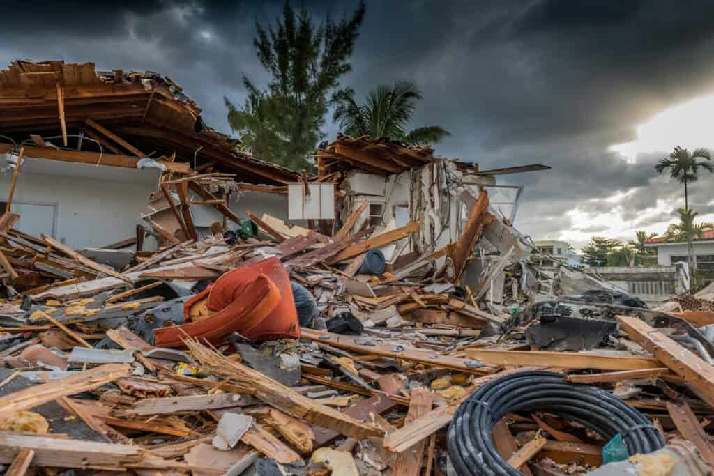 A severely damaged house with debris scattered everywhere, indicating the aftermath of a natural disaster. Dark clouds loom overhead, adding to the sense of devastation.