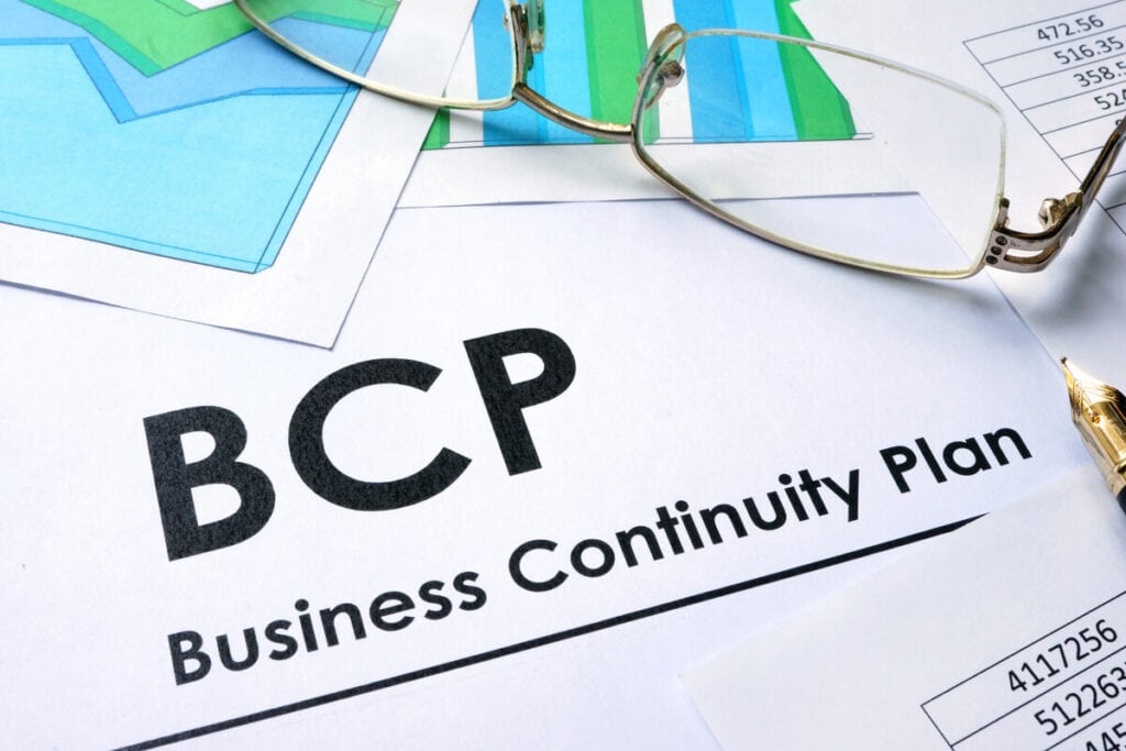 Close-up of a document titled "BCP Business Continuity Plan" with a pair of glasses and financial charts in the background, highlighting the importance of planning for business continuity.