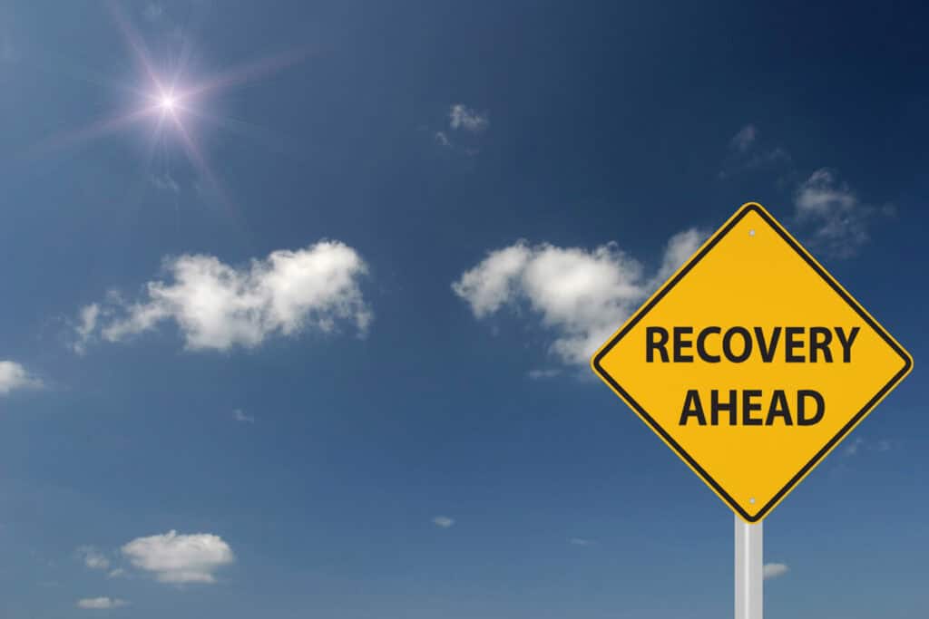 A bright blue sky with a few scattered white clouds and a shining sun in the background. In the foreground, a yellow road sign reads "RECOVERY AHEAD," symbolizing hope and progress after a disaster.