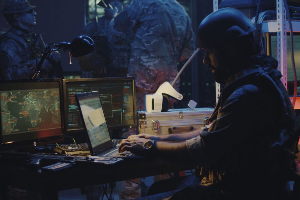 A military officer in tactical gear is working on a laptop, surrounded by multiple screens displaying maps and data. Other personnel in uniform are in the background, indicating a high-tech command center focused on disaster recovery operations.